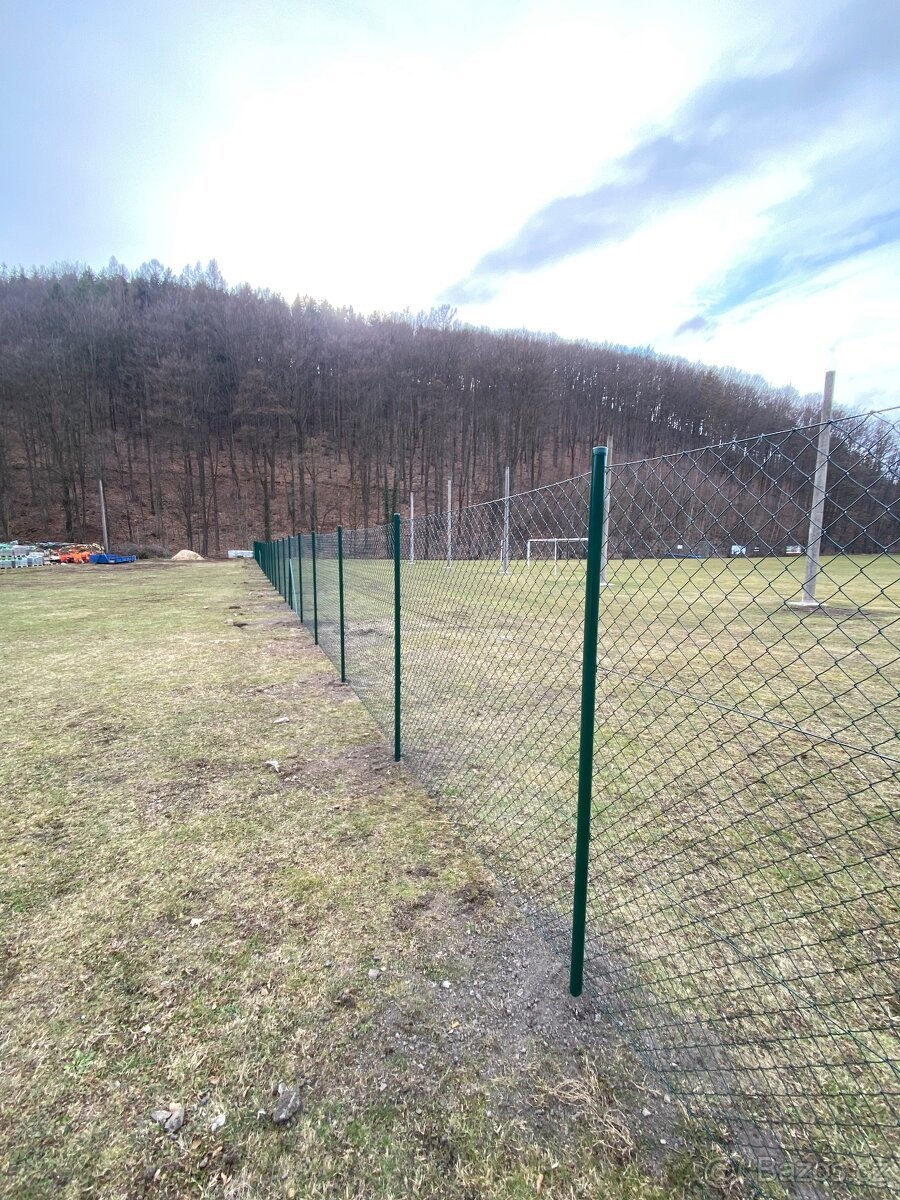 Pletivové oplocení se zelenými sloupky kolem sportovního hřiště pod zalesněným kopcem